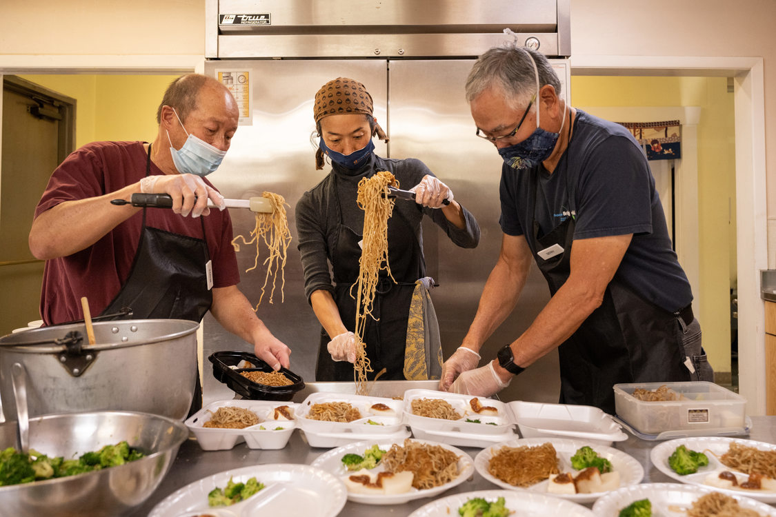 Three cooks place long strings of noodles in to-go food containers